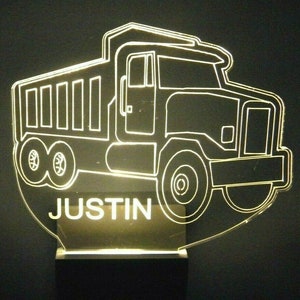 May include: A white outline of a dump truck with the name "JUSTIN" underneath. The truck is illuminated from behind, creating a glowing effect.