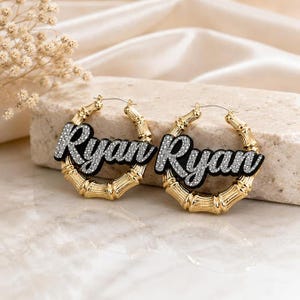 May include: A pair of gold-toned bamboo hoop earrings featuring the name Ryan in a cursive, silver-toned glitter script layered over a black background. The earrings have a circular bamboo-textured design and standard wire earring posts.