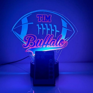 May include: A blue and pink 3D printed night light shaped like a football with the text "TIM Buffalo" on it. The night light is on a black base with a remote control.