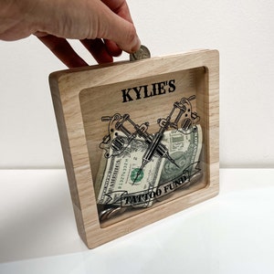 May include: A wooden shadow box with a clear front, labeled "KYLIE'S" and "TATTOO FUND". The box contains a dollar bill, coins, and a graphic of two tattoo machines. A hand is dropping a coin into the slot.