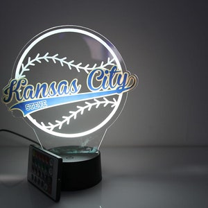 May include: A glowing baseball-themed night light. The design features a baseball with stitching and the words "Kansas City" in blue and gold, with the name "Steve" below. Includes a remote control.