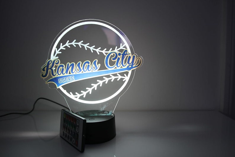 May include: A glowing baseball-themed night light. The design features a baseball with stitching and the words "Kansas City" in blue and gold, with the name "Steve" below. Includes a remote control.