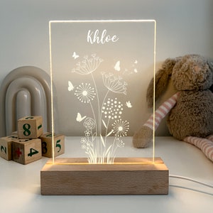 May include: A white acrylic night light with a wooden base. The light features a dandelion design with butterflies and the name Khloe.