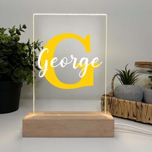 May include: A personalized night light with the name "George" in white script over a yellow "G" on a clear acrylic panel. The panel is set in a light-colored wooden base, illuminated by a warm glow. A decorative item for a bedroom or nursery.