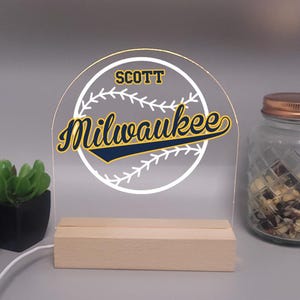 May include: A clear acrylic baseball-themed night light with the name "Scott" and "Milwaukee" in navy and gold lettering. The light is set on a wooden base and has a baseball design. A small plant and a jar are in the background.