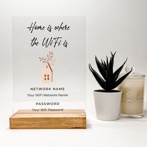 May include: A clear acrylic sign with a house illustration and the text "Home is where the WiFi is". The sign also includes the text "NETWORK NAME" and "PASSWORD" with placeholders for the network name and password. The sign is on a wooden stand.