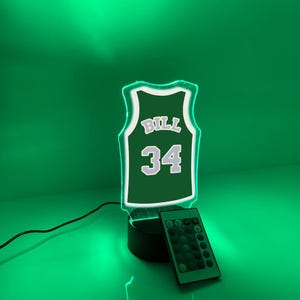 May include: A green and white LED night light shaped like a basketball jersey with the name "BILL" and the number "34". The light sits on a black base and includes a remote control.