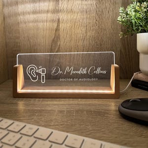 May include: A lighted desk sign with a wooden base and a clear acrylic panel. The panel displays an ear and audiometer graphic, along with the text "Dr. Meredith Collins, DOCTOR OF AUDIOLOGY". The sign is illuminated with a warm glow.