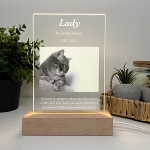 Könnte beinhalten: Eine Gedenk-LED-Leuchte mit Holzsockel. Die transparente Acrylplatte zeigt ein Graustufenfoto einer Katze namens Lady mit dem Text "In Loving Memory 2007-2021". Der Sockel hat einen warmen Schein, und der Text "No home is complete..." ist ebenfalls sichtbar.