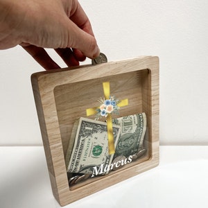 Könnte beinhalten: Eine hölzerne Spardose mit einer Glasfront, die ein goldenes Kreuz mit floralen Akzenten und den Namen "Marcus" unten eingraviert zeigt. Die Dose ist mit Münzen und Dollarnoten gefüllt.