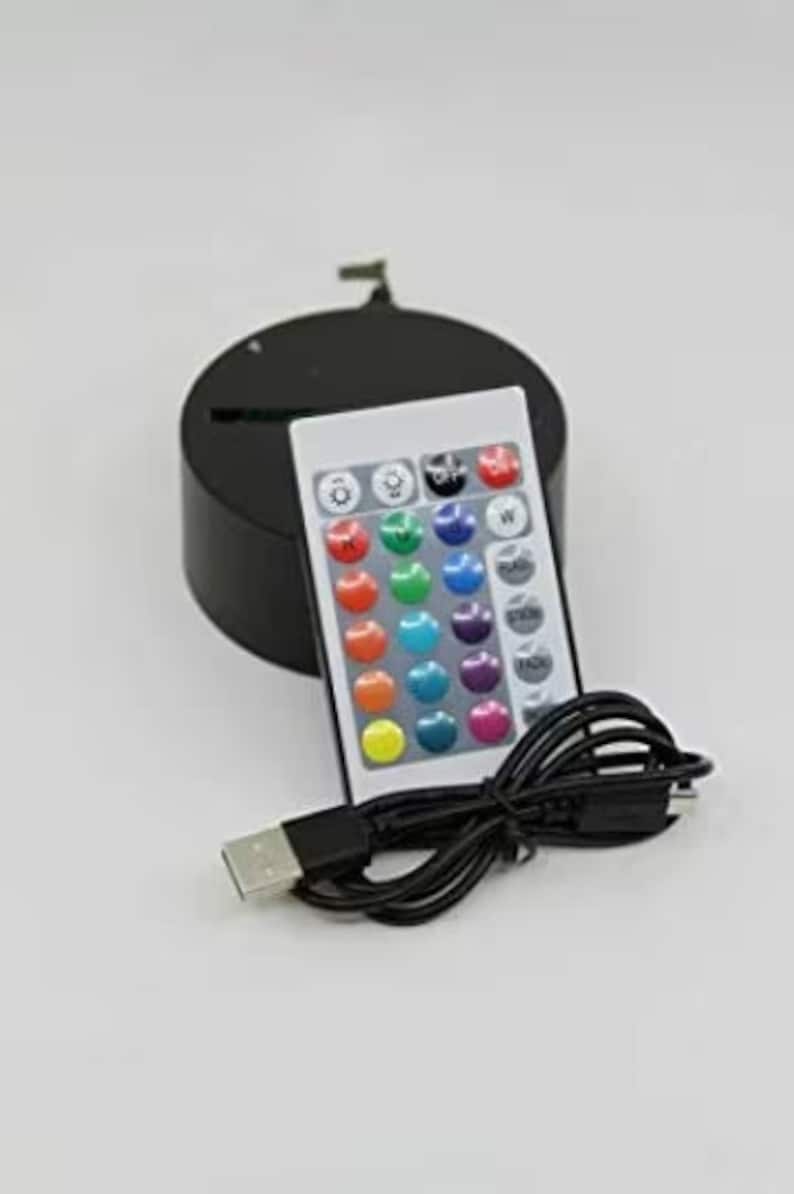 May include: A black LED light base with a remote control and USB cable. The remote has multiple color options and control buttons. The base is round and has a slot for an item to be displayed.