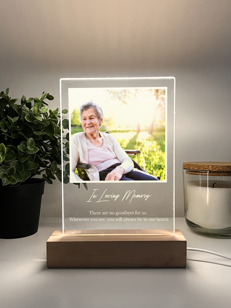 May include: A white acrylic photo frame with a wooden base. The frame has a photo of an older woman sitting on a bench in a park. The text "In Loving Memory" is written on the frame, along with the quote "There are no goodbyes for us. Whenever you are, you will always be in our hearts."