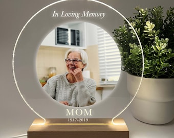 May include: A white acrylic circle with a photo of a woman in the centre. The circle is lit from behind and has the text "In Loving Memory" at the top and "MOM 1947-2019" at the bottom. The circle is sitting on a wooden base.