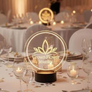 May include: A circular acrylic sign with a gold-colored logo featuring a stylized flower and the text Crown Flower Jewelry, mounted on a black cylindrical base, positioned on a white tablecloth with wine glasses and lit candles.