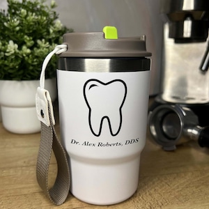 May include: White travel mug featuring a black tooth graphic and the text "Dr. Alex Roberts, DDS." The mug has a grey lid with a green button and a grey strap. A small plant and coffee machine are in the background.
