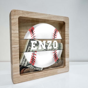 May include: Wooden baseball-themed money box with a clear front panel. The box has a baseball inside and the name "ENZO" printed on the front.