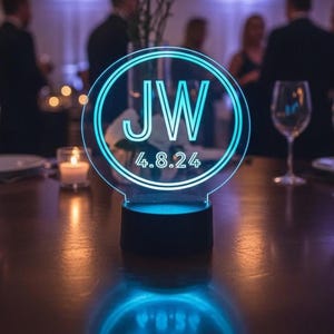 Puede incluir: Un letrero circular de acrílico con un efecto de luz cian brillante, que presenta las letras JW y la fecha 4.8.24 centradas dentro de anillos de doble línea. El letrero está montado sobre una base cilíndrica negra que ilumina el panel.