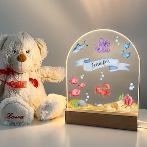 Puede incluir: Una lámpara de noche decorativa con base de madera y panel acrílico arqueado. El panel presenta una escena submarina con peces, un cangrejo y el nombre "Jennifer" en una pancarta. Un oso de peluche con una cinta roja está a la izquierda.