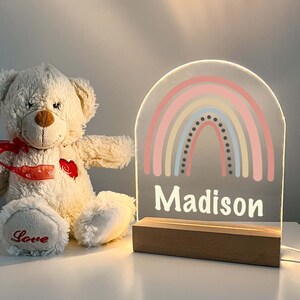 May include: A white acrylic night light with a rainbow design and the name "Madison" on the bottom. The night light is on a wooden base and is lit up.