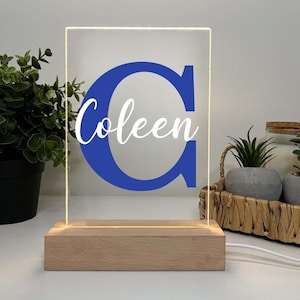 May include: A personalized LED night light featuring a blue letter "C" and the name "Coleen" in white script. The light sits on a rectangular wooden base and is plugged in with a white cord. The light is on.