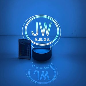 Puede incluir: Una luz nocturna LED blanca con un panel acrílico circular que presenta las iniciales "JW" y la fecha "4.8.24" en un diseño de doble círculo. La luz está sobre una base negra y viene con un control remoto.