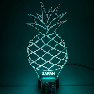 Puede incluir: Una luz nocturna en forma de piña de color azul turquesa con un mando a distancia. La luz tiene un nombre personalizado, "SARAH", en la base.