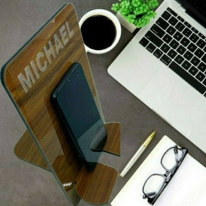 Könnte beinhalten: Ein Holz-Telefonständer mit einer personalisierten Namensschilder, auf dem "MICHAEL" steht. Der Ständer ist braun mit einer Holzmaserung und hat einen Schlitz für ein Telefon. Ein schwarzes Smartphone befindet sich im Schlitz.
