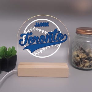 May include: A clear acrylic baseball-themed night light with the name "Jason" and the word "Toronto" in blue script. The light sits on a wooden base and has a white baseball design. A small plant and a jar are in the background.