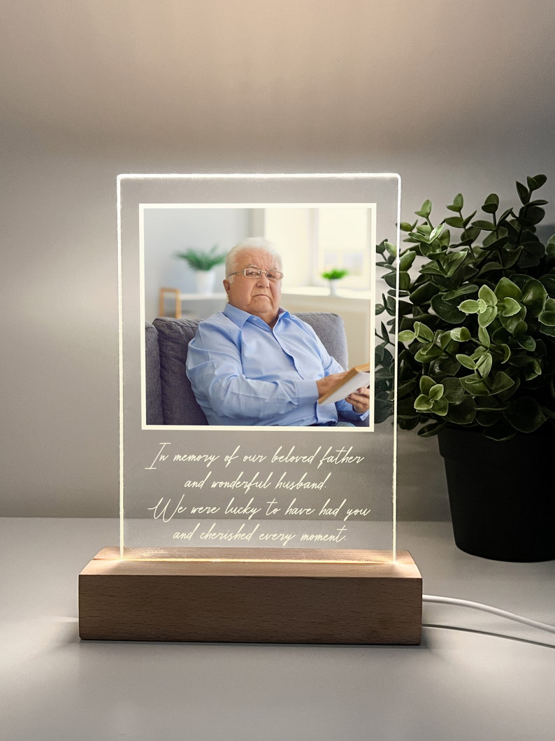 May include: A white acrylic photo frame with a wooden base. The frame has a photo of an older man in a blue shirt. The frame has a message that reads "In memory of our beloved father and wonderful husband. We were lucky to have had you and cherished every moment."