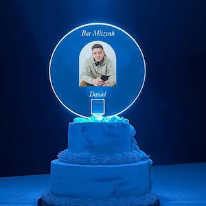 May include: A two-tiered blue cake topped with a circular illuminated sign. The sign features a photo of a young man, the words "Bar Mitzvah" and the name "Daniel". The cake is decorated with white frosting.