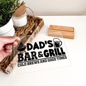 May include: A clear acrylic sign with black text that reads "Dad's Bar & Grill Cold Brews and Good Times". The sign features a grill and a beer mug.