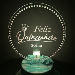 May include: A round, clear acrylic cake topper with a white crown and the text "Feliz Quinceañera Sofia" in a decorative script font. The topper is lit from behind by a small LED light.
