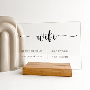 Könnte beinhalten: Ein klares Acrylschild mit dem Wort "wifi" in Kursivschrift. Das Schild hat zwei Abschnitte, einer mit der Bezeichnung "Netzwerkname" und der andere mit der Bezeichnung "Passwort". Das Schild steht auf einem Holzständer.