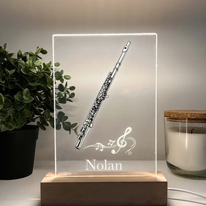 Peut inclure: Une veilleuse en acrylique blanche avec une base en bois. La lumière présente une illustration en noir et blanc d'une flûte avec des notes de musique et le nom "Nolan".