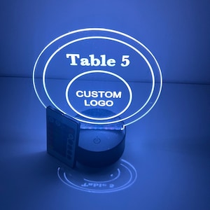 May include: A white LED table number sign with a clear acrylic base. The sign displays "Table 5" in a circle with a smaller circle inside that says "CUSTOM LOGO". The sign is illuminated with a blue light and has a remote control.