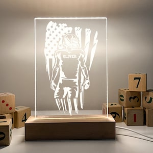 Könnte beinhalten: Eine weiße Silhouette eines American-Football-Spielers mit dem Namen "OLIVER" auf dem Trikot, der vor einem amerikanischen Flaggenmuster steht. Das Bild wird von hinten beleuchtet, wodurch ein leuchtender Effekt entsteht. Das Bild befindet sich auf einem Holzsockel.