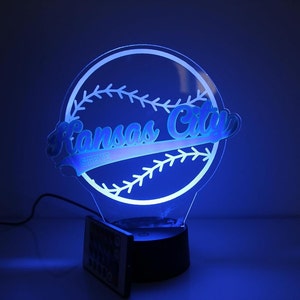 May include: A blue-lit baseball-themed night light. The design features the words "Kansas City" in a script font, with "STEVE" below, set against a baseball outline. The light has a black base and includes a remote control.