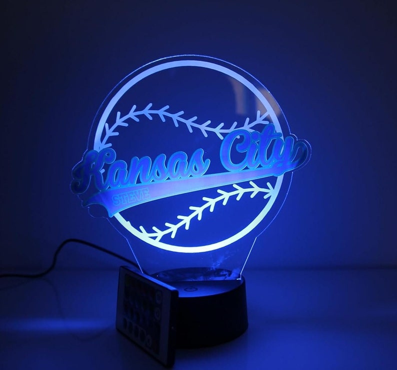 May include: A blue-lit baseball-themed night light. The design features the words "Kansas City" in a script font, with "STEVE" below, set against a baseball outline. The light has a black base and includes a remote control.