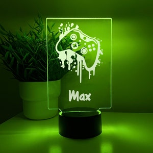 Könnte beinhalten: Ein klares Acryl-Nachtlicht mit einer weißen Silhouette eines Videospiel-Controllers und dem Namen Max. Das Licht befindet sich auf einem schwarzen Sockel und wird mit grünem Licht beleuchtet.