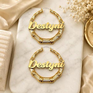 May include: A pair of gold-toned hoop earrings featuring a bamboo-inspired texture. Each earring has a central horizontal nameplate displaying the name Destyni in a cursive script font. The earrings are circular with a polished metallic finish.