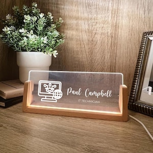 May include: A desk nameplate with a wooden base and a clear acrylic panel. The panel is illuminated and displays the name "Paul Campbell" and the title "IT TECHNICIAN" in white. A computer graphic is on the left.