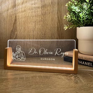 Può includere: Targhetta da scrivania illuminata con base in legno. Il pannello in acrilico trasparente mostra il nome "Dr. Olivia Reyes" e il titolo "SURGEON" in elegante scrittura. Un'illustrazione di un chirurgo è a sinistra.
