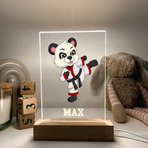 Puede incluir: Un personaje de dibujos animados de panda blanco y negro que lleva un uniforme de karate blanco con un cinturón rojo, dando una patada en el aire. La imagen está en un panel de acrílico transparente con una base de madera. El nombre "MAX" está impreso en amarillo debajo del dibujo animado.