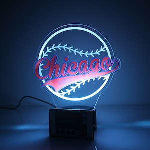 Może przedstawiać: Biało-niebieska lampka nocna LED w kształcie baseballu z napisem "Chicago" w czerwonych i niebieskich literach. Nazwa "Larry" jest napisana mniejszymi literami pod słowem "Chicago".