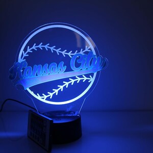 May include: A blue-lit acrylic baseball-themed lamp featuring the words "Kansas City" in a stylized font. The lamp is shaped like a baseball with stitching details and sits on a black base. Includes a remote control.