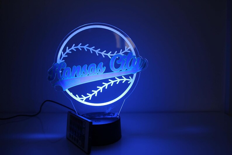 May include: A blue-lit acrylic baseball-themed lamp featuring the words "Kansas City" in a stylized font. The lamp is shaped like a baseball with stitching details and sits on a black base. Includes a remote control.