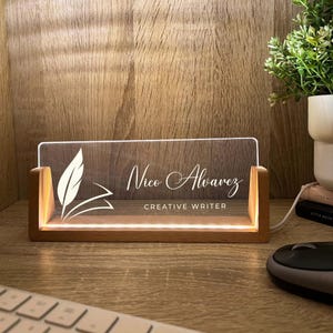 May include: A lighted desk nameplate with a wooden base. The clear acrylic panel displays the name "Nico Alvarez" and the title "CREATIVE WRITER" in white script. A feather and book design is on the left. The nameplate is illuminated by a warm light.