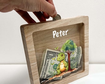 May include: Wooden coin bank with a clear front panel. The bank features a cartoon dinosaur and a tree. The name "Peter" is written on the front of the bank.