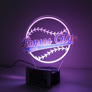 May include: A baseball-themed LED night light with the words "Kansas City" in a stylized font, and the name "Steve" below. The light is white and blue, with a black base and a remote control.