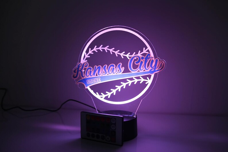 May include: A baseball-themed LED night light with the words "Kansas City" in a stylized font, and the name "Steve" below. The light is white and blue, with a black base and a remote control.